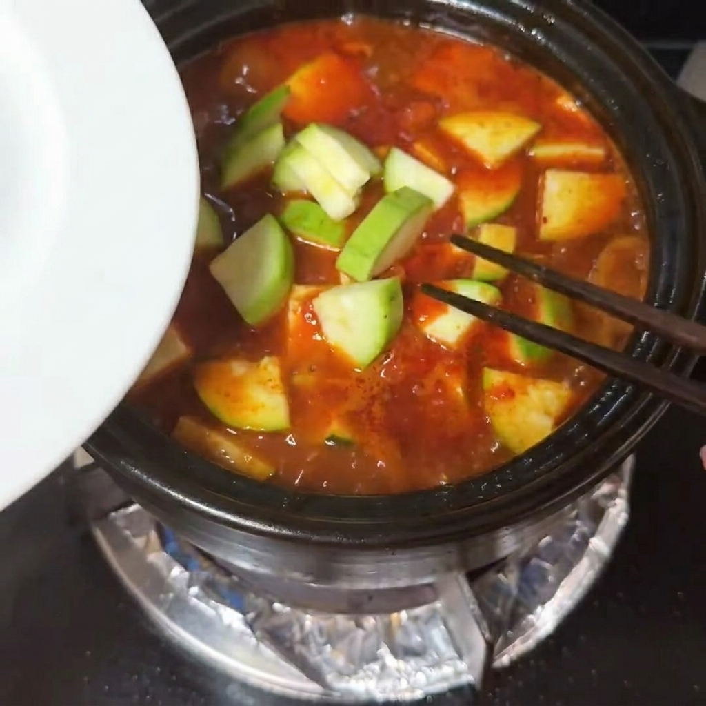 Zucchinischeiben und Stücke aus weichem Seidentofu werden in die sprudelnde rote Suppe gegeben.