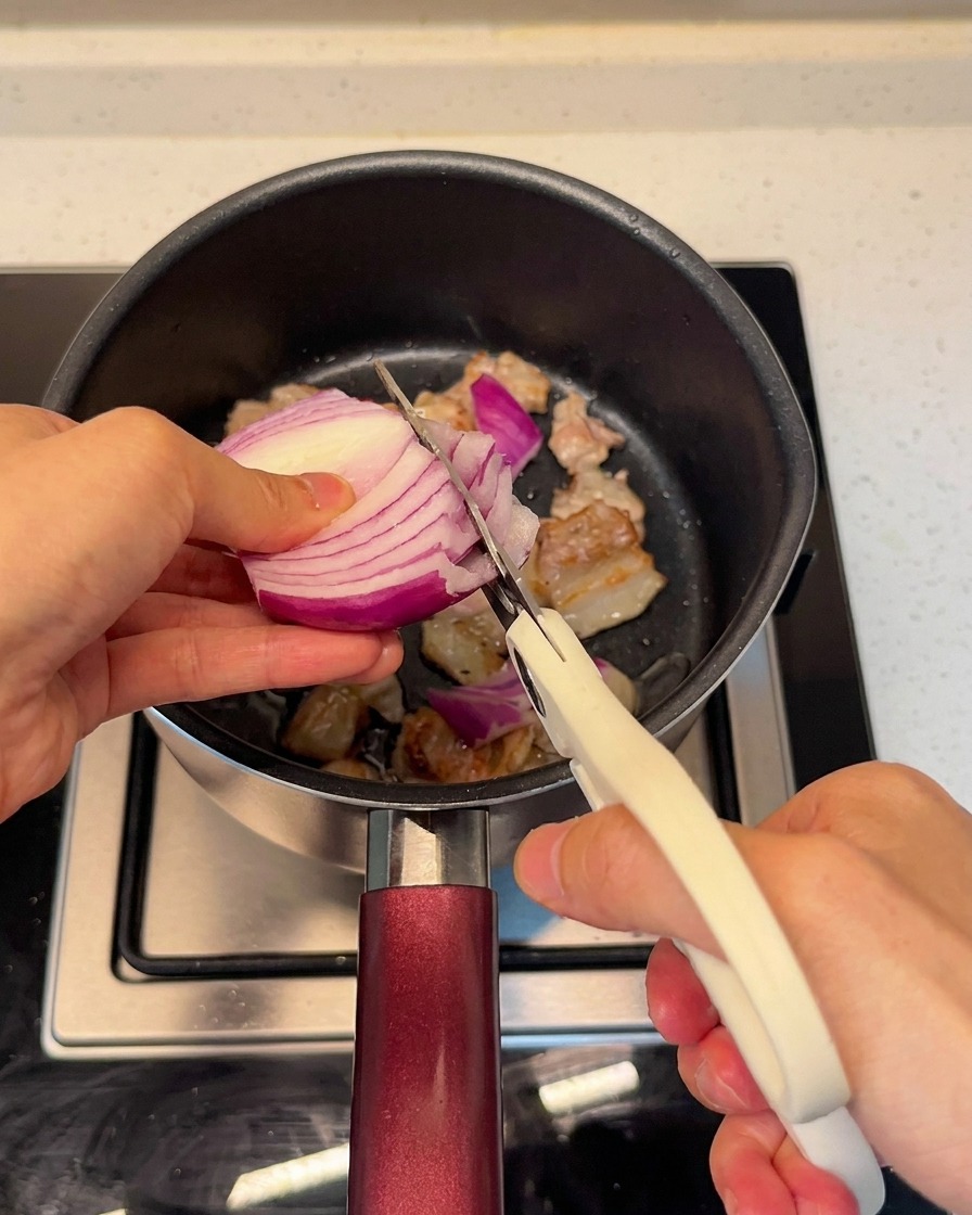 Frische rote Zwiebeln werden mit einer Küchenschere direkt in den Topf zum Schweinebauch geschnitten.
