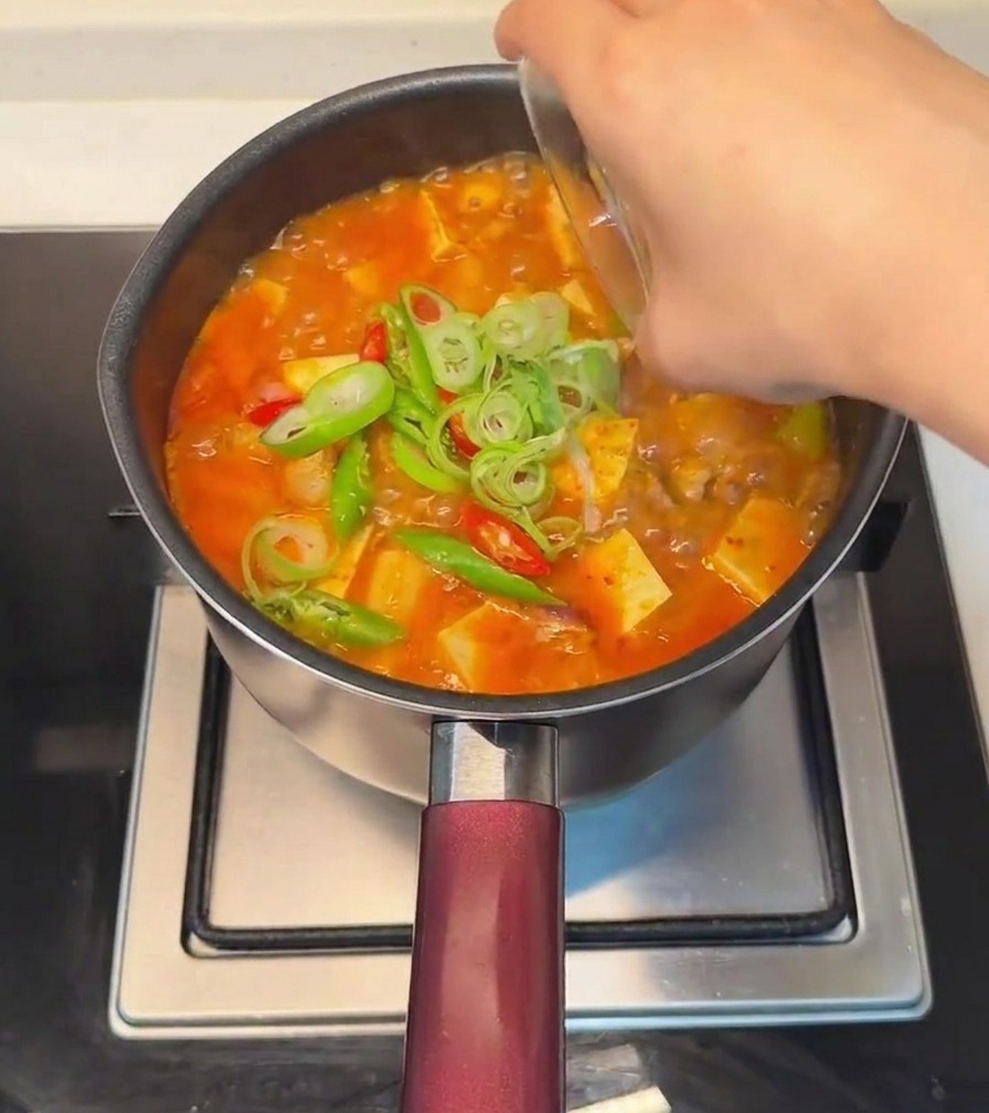 Eine Hand streut geschnittene Frühlingszwiebeln sowie frische rote und grüne Chilischoten über den fertigen Tofu-Eintopf.