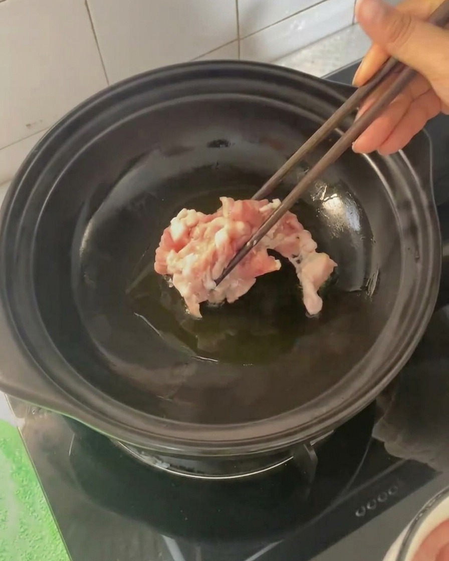 Schweinebauchscheiben werden mit Stäbchen in den heißen Tontopf gegeben.