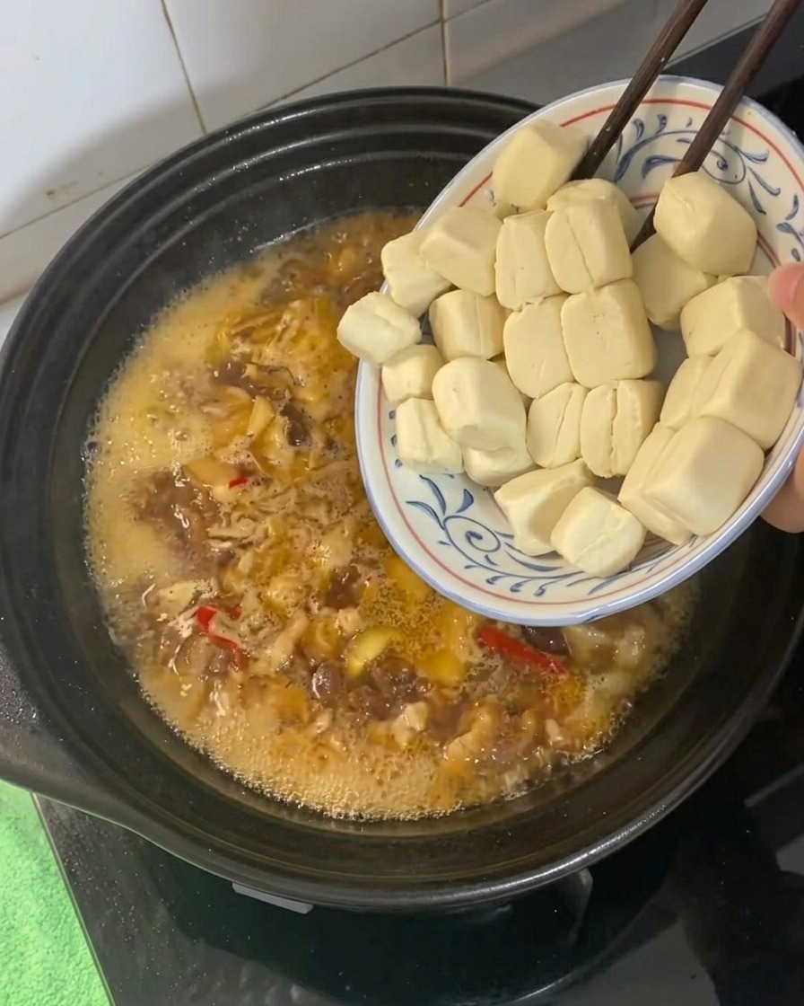 Kleine Tofu-Würfel werden in eine sprudelnde, goldbraune Schmorflüssigkeit in einem Tontopf gegeben.