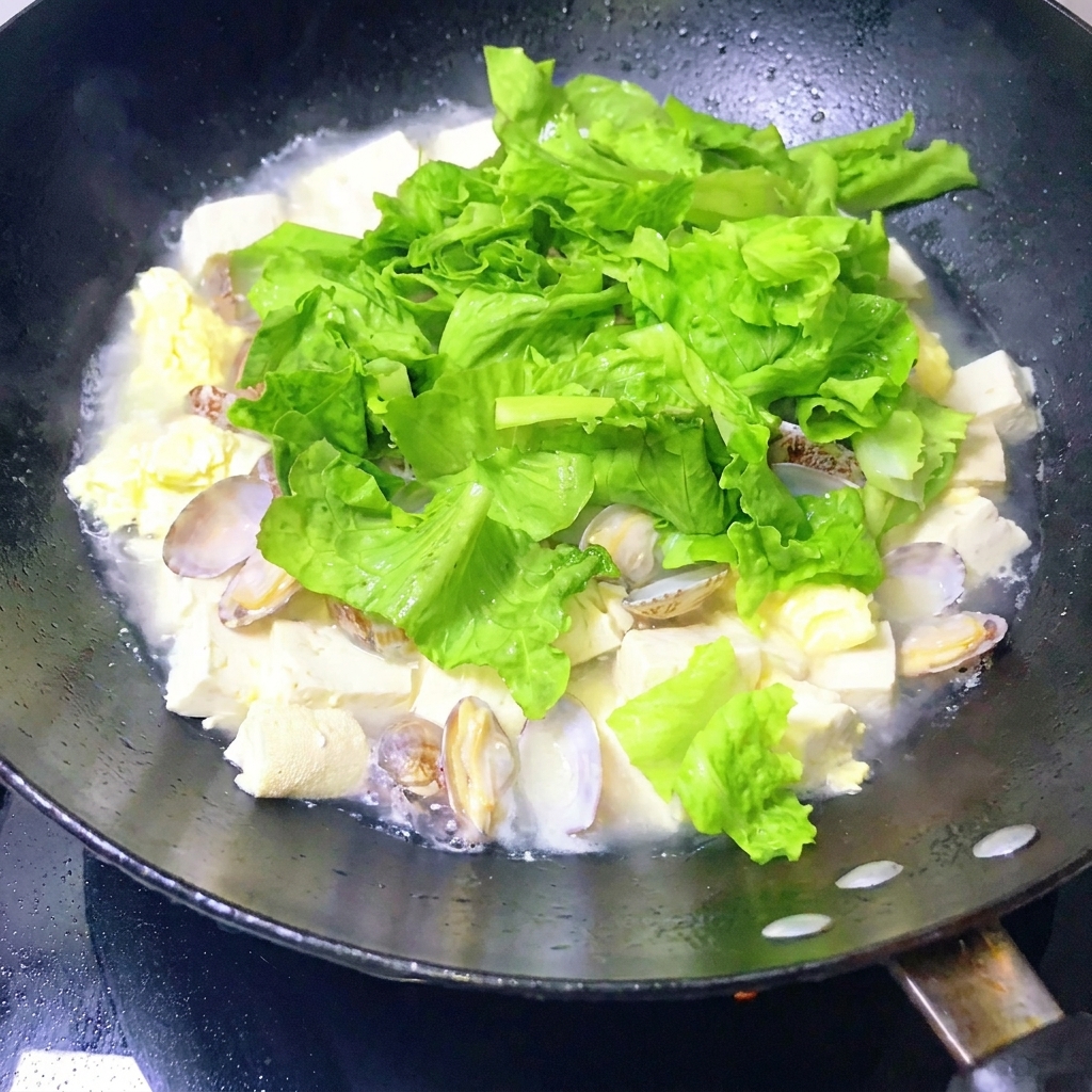Eine Pfanne mit fertigem Muschel-Tofu-Eintopf wird vorsichtig mit einer Schöpfkelle umgerührt.