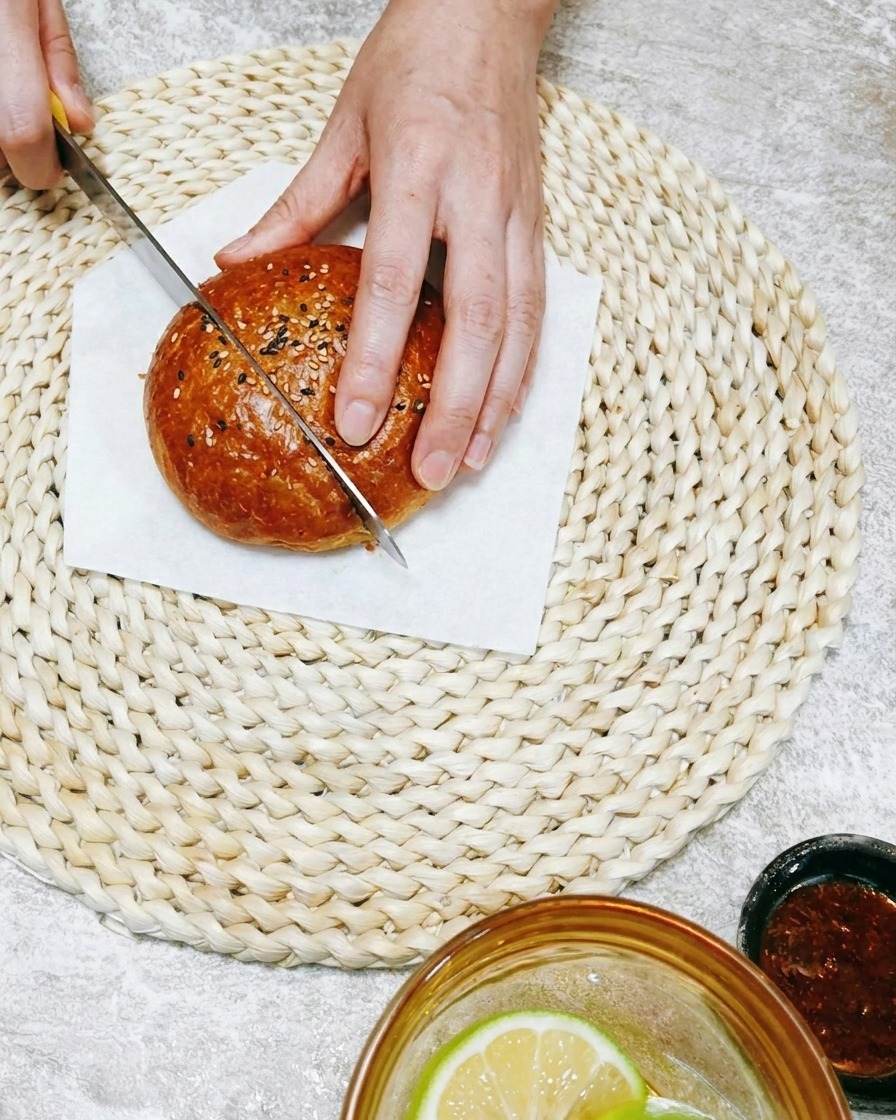 Nahaufnahme einer Hand, die mit einem Messer ein weiches Burgerbrötchen auf einer gewebten Unterlage schneidet.