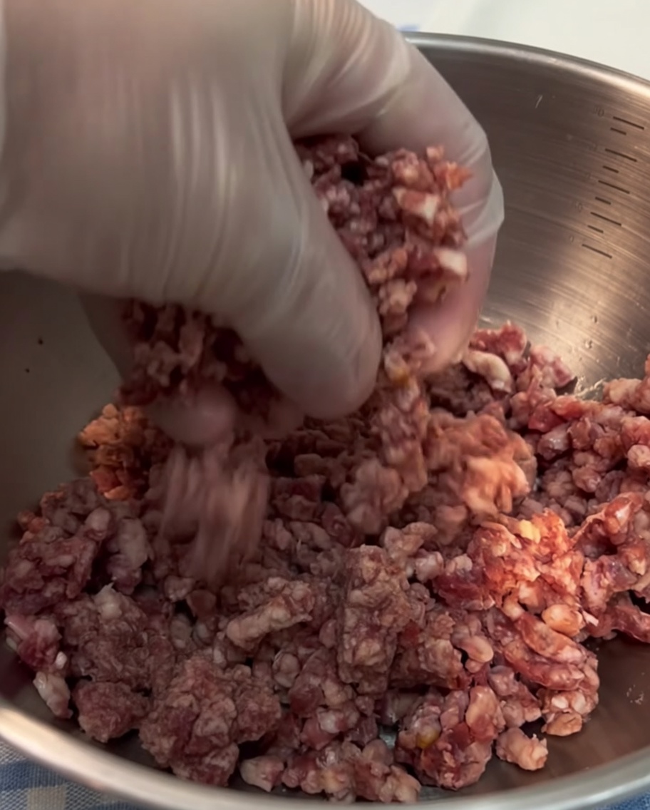 Nahaufnahme einer Hand in einem Einweghandschuh, die frisches Rinderhackfleisch zu einer kompakten Kugel formt.