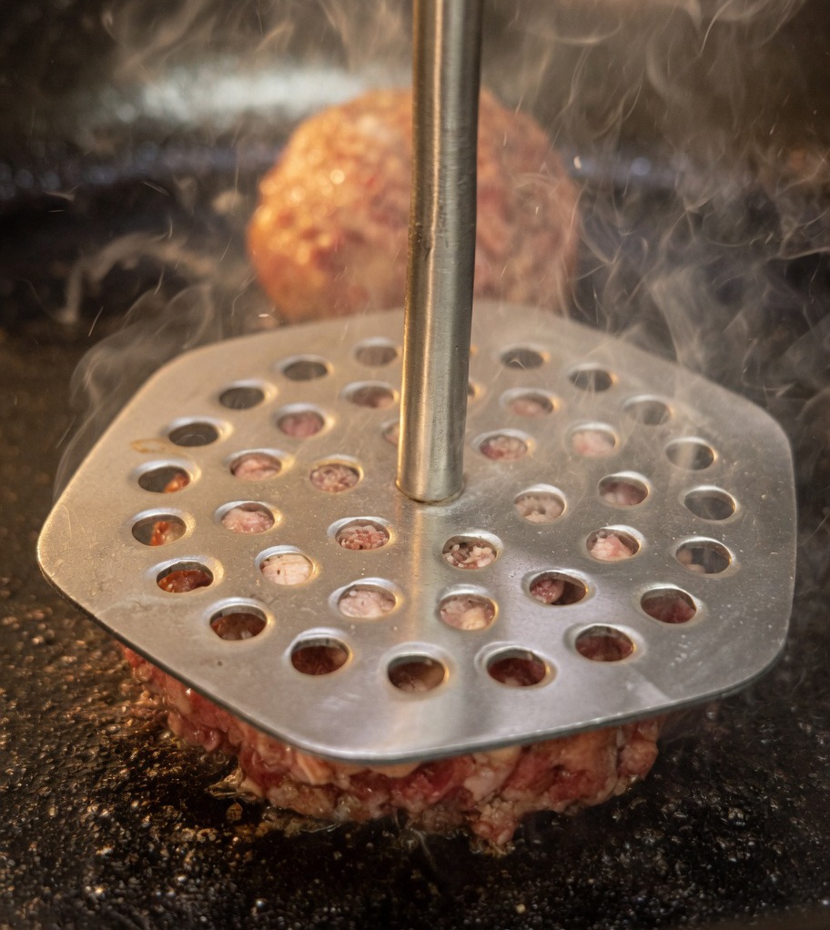 Eine Fleischpresse aus Metall drückt die Rindfleischbällchen in einer heißen Pfanne flach.