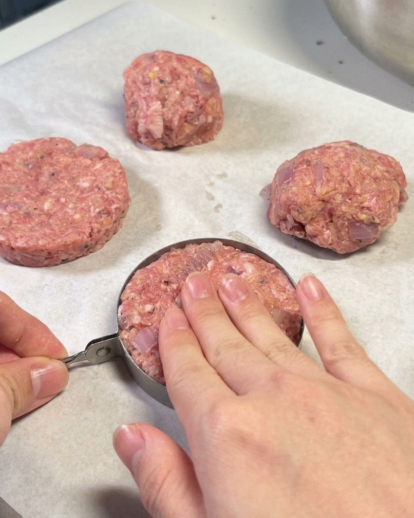 Hände benutzen einen Metallring, um ein Hamburger-Patty auf Backpapier zu formen.