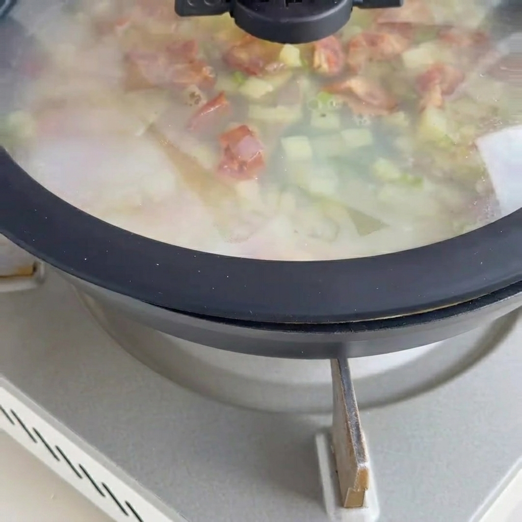 Cremige Muschelsuppe simmert in einer Pfanne mit Muschelfleisch und Speckstücken.