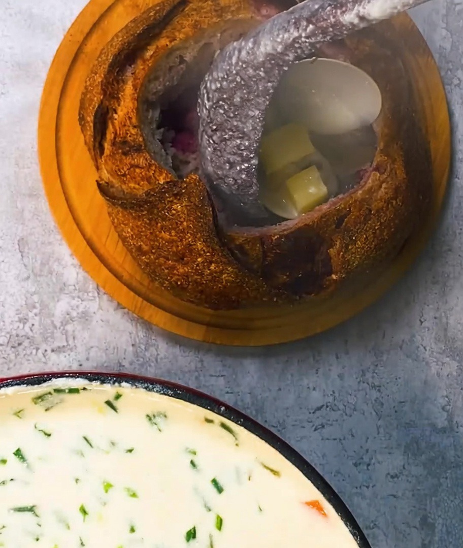 Reiche, cremige Muschelsuppe wird aus einer Schöpfkelle in einen rustikalen Brotlaib gefüllt und mit knusprigen Speckstücken garniert.