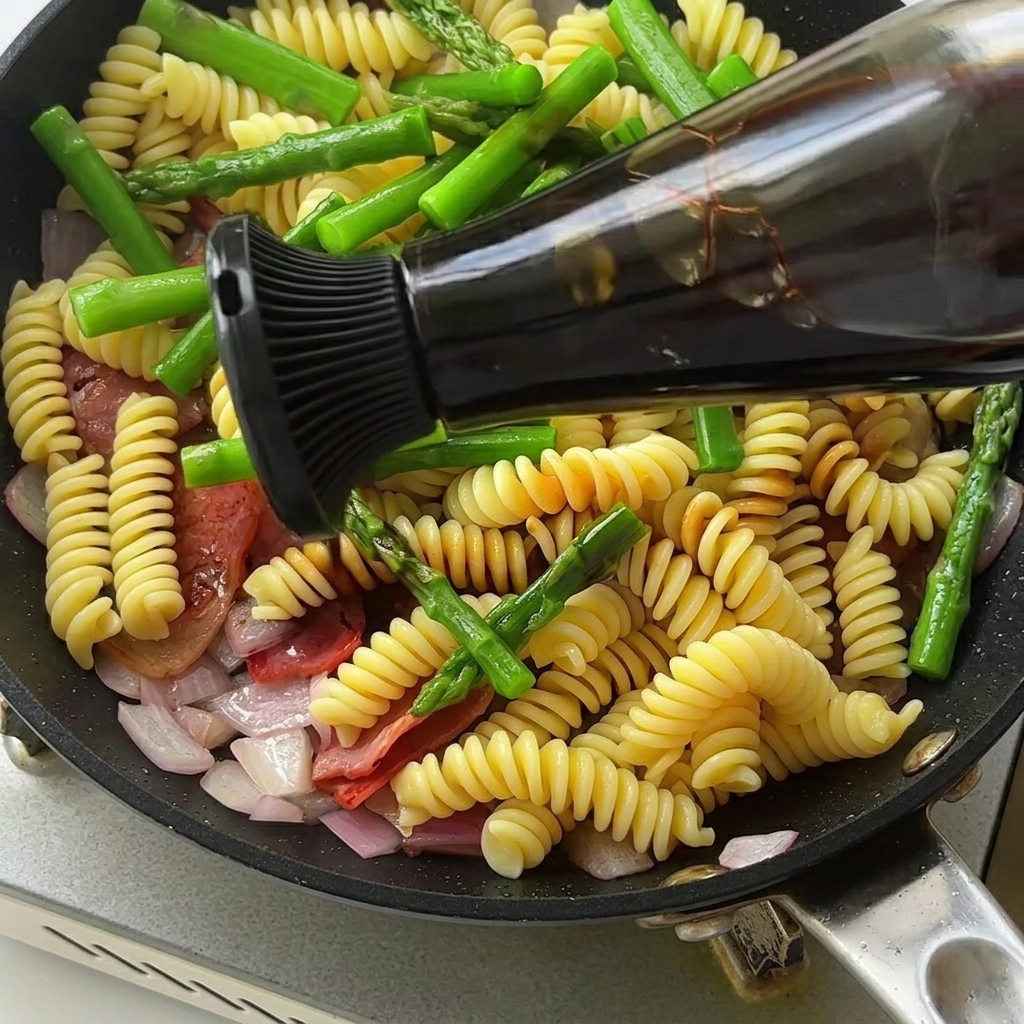 Nahaufnahme einer Pfanne mit Fusilli, Spargel und Speck, die mit einer dunklen, reichhaltigen Sauce verfeinert wird.