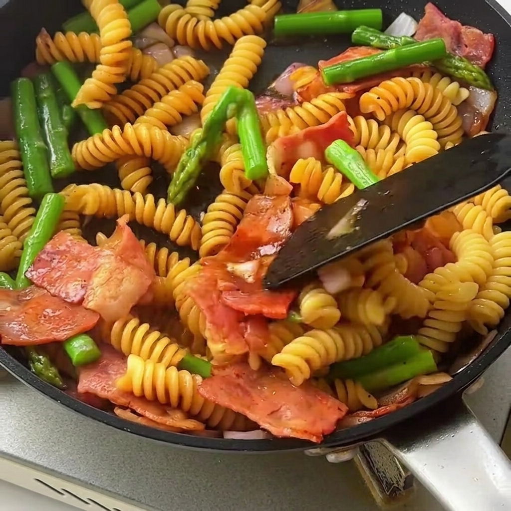 Ein Pfannenwender vermengt die glänzende Pasta, den Spargel und Speck in einer großen Pfanne.