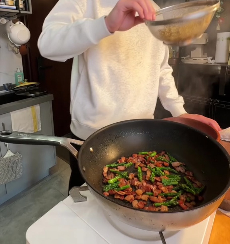 Gekochte Penne-Pasta wird in eine Pfanne mit knusprigem Speck und angebratenen Spargelspitzen gegeben.