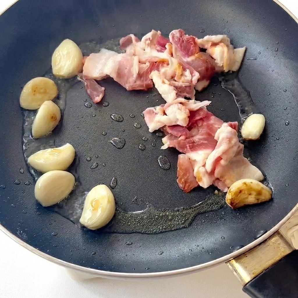 Speck brät in einer Pfanne neben gebräunten Knoblauchzehen.
