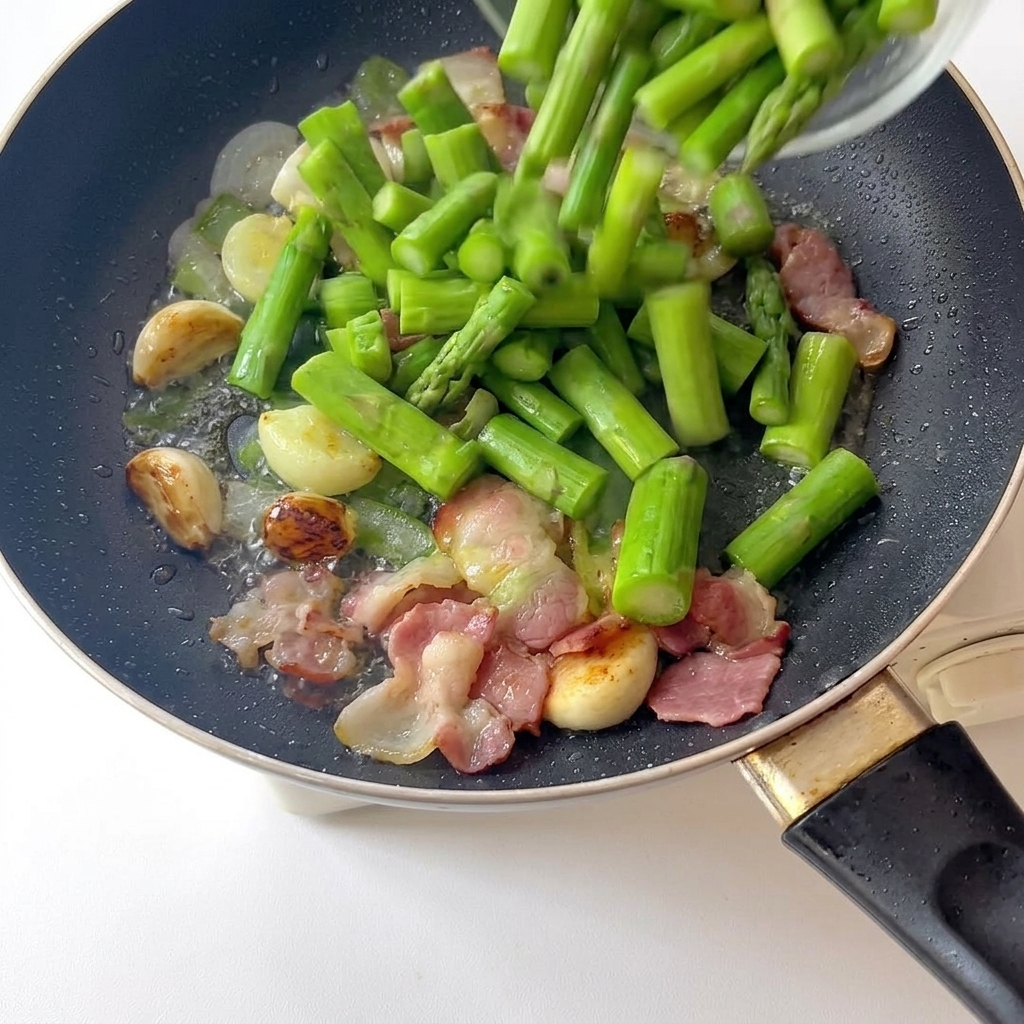 Frisch blanchierte, hellgrüne Spargelstücke werden in eine Pfanne mit Knoblauch und Speck gegeben.