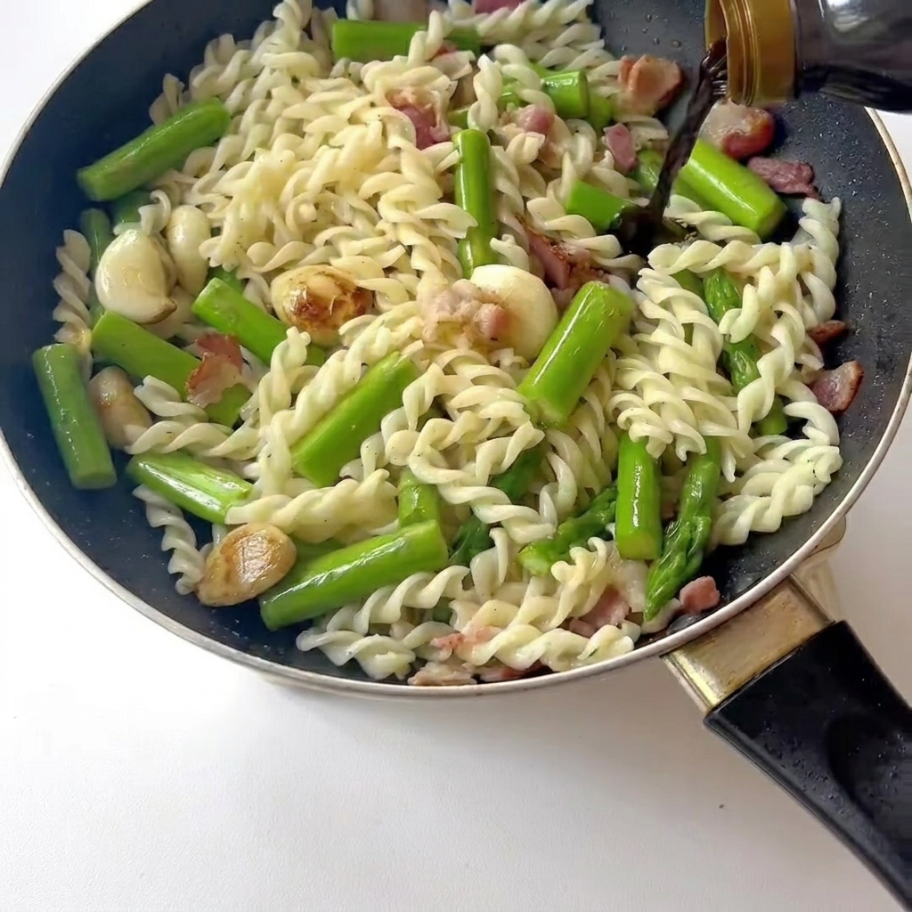 Nahaufnahme beim Umrühren von Pasta, Speck und Spargel, während helle Sojasauce darüber gegeben wird.