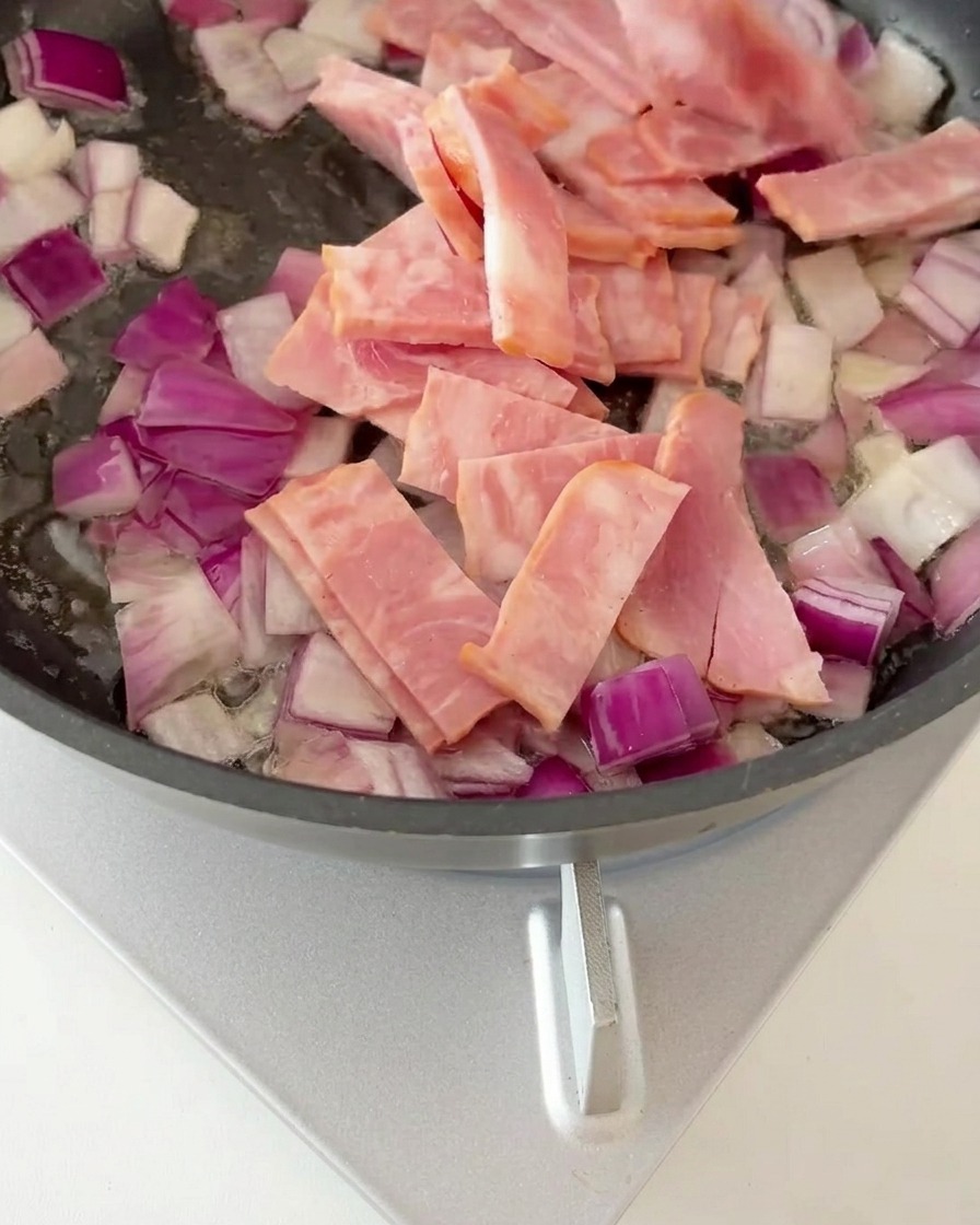Speckstücke werden mit angeschwitzten Zwiebeln in einer Pfanne goldbraun gebraten.