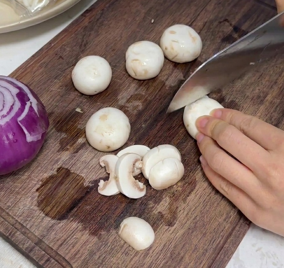 Frische weiße Champignons werden auf einem Holzschneidebrett in gleichmäßige Stücke geschnitten.