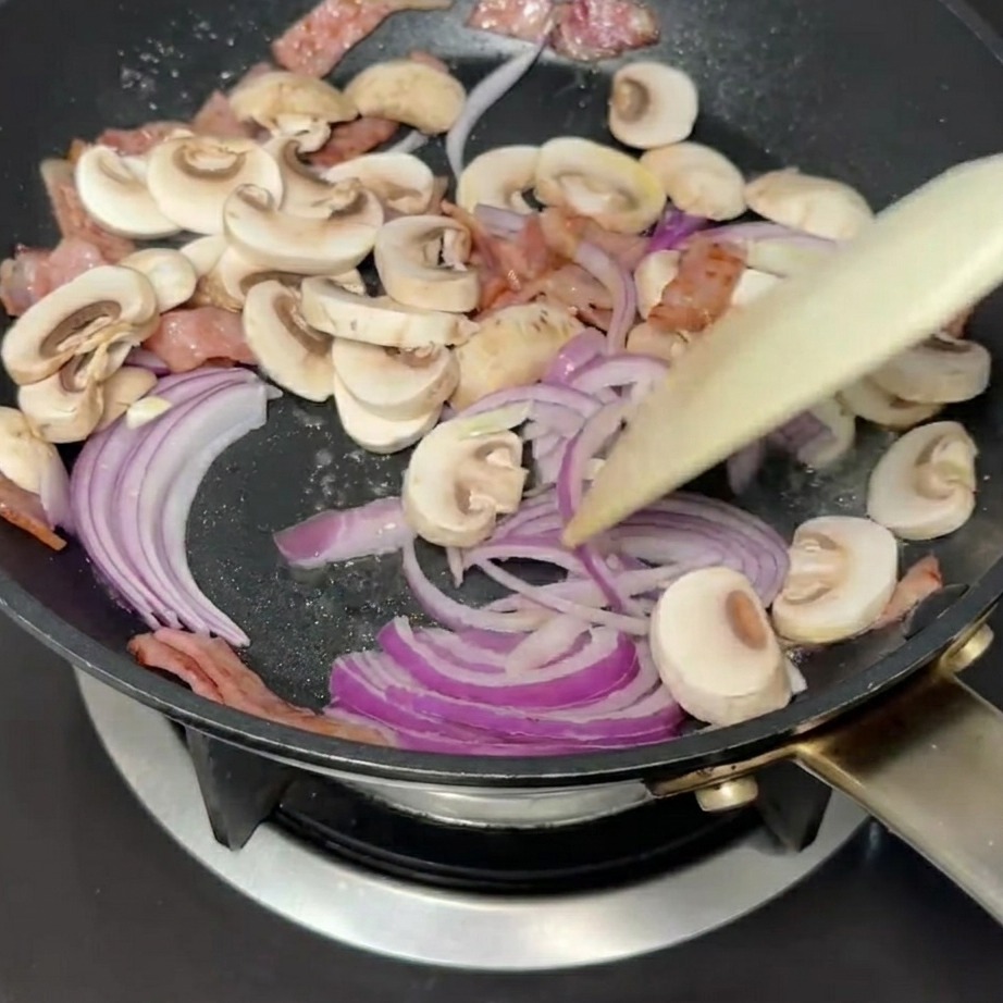 Geschnittene rote Zwiebeln und weiße Champignons werden in einer Pfanne mit Speck angebraten.