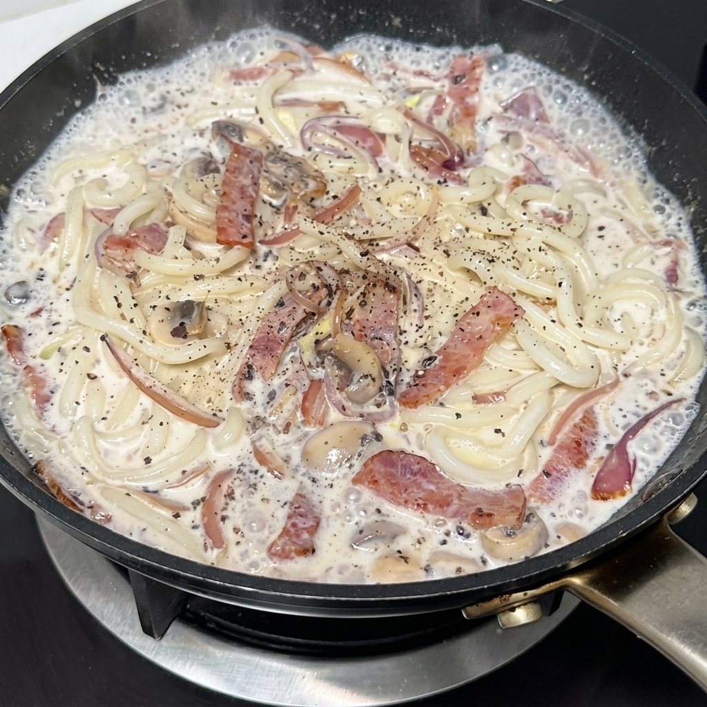 Eine Pfanne mit cremigen Udon-Nudeln, Speck und Pilzen wird mit schwarzem Pfeffer gewürzt.