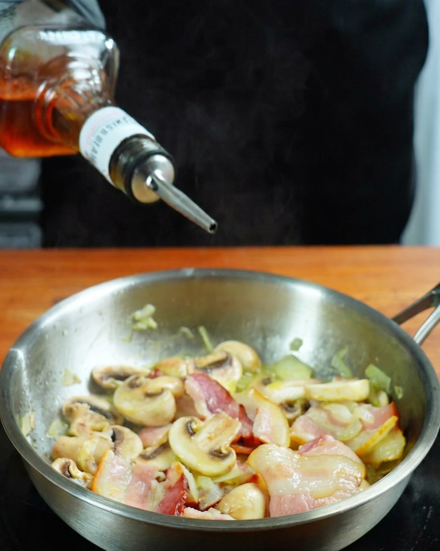 Whisky wird aus einer Flasche in eine Pfanne mit Speck und Pilzen gegossen.