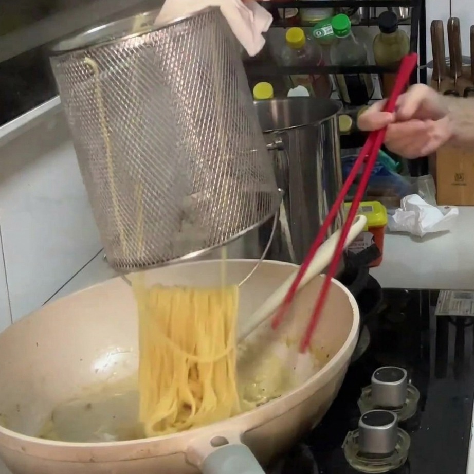 Gekochte Spaghetti werden aus einem Sieb in eine Pfanne mit cremiger Pilzsauce gegeben.