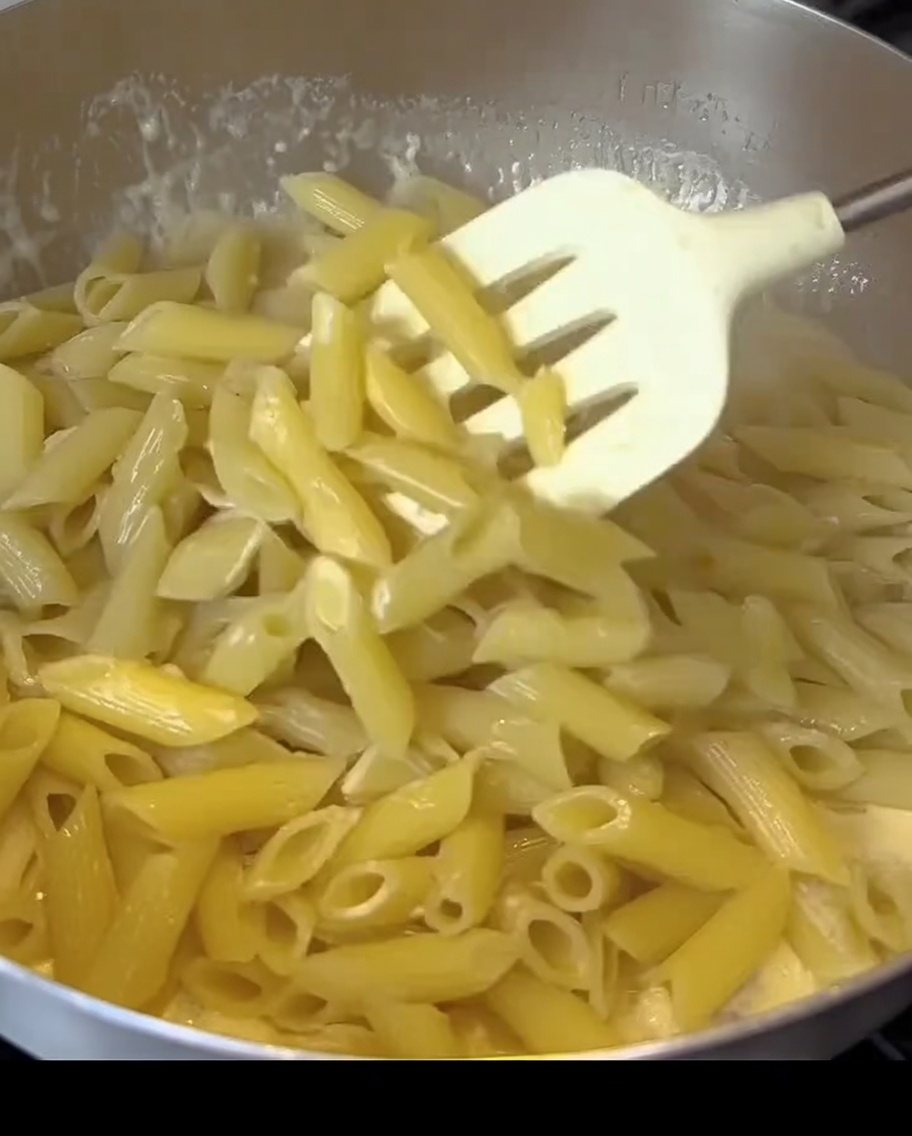 Penne-Pasta, die in einer cremigen weißen Sauce mit Speckstücken in einer Pfanne vermengt wird.