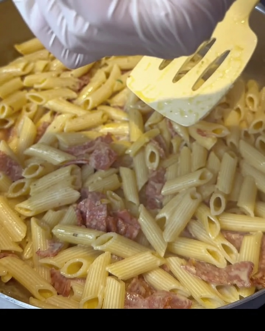 Handschuhe, die Gewürze über eine Pfanne mit cremiger Speck-Pasta streuen.