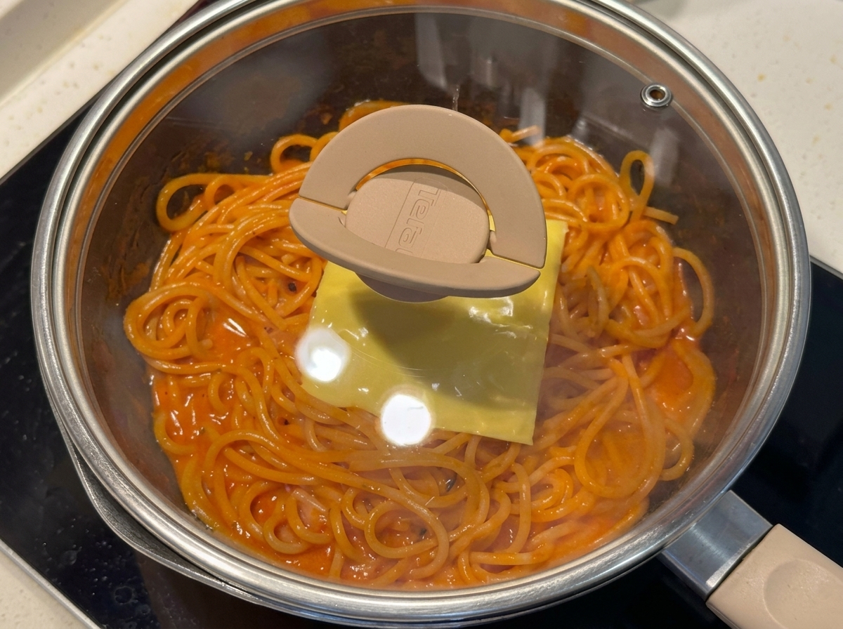 Eine Pfanne mit Spaghetti, die mit einer Scheibe Käse belegt und mit einem Glasdeckel abgedeckt ist.