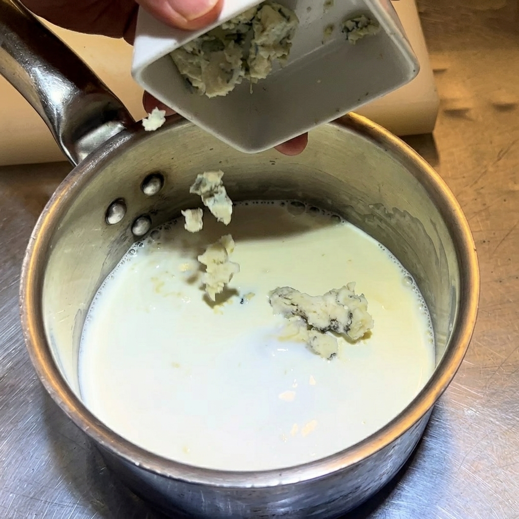 Zerbröckelter Blauschimmelkäse wird in einen Topf mit Milch und Sahne gegeben.