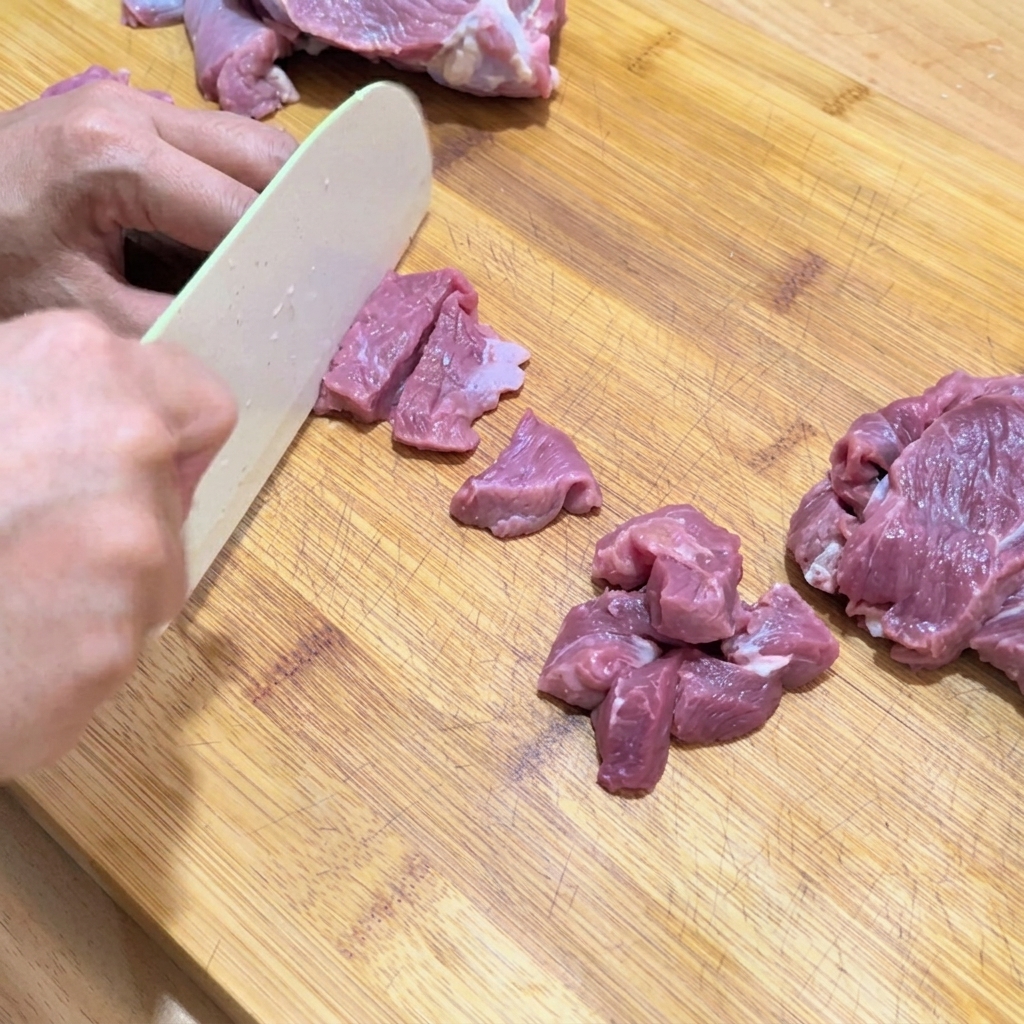 Hände schneiden rohes Rindfleisch auf einem Holzbrett in kleine, gleichmäßige Würfel.