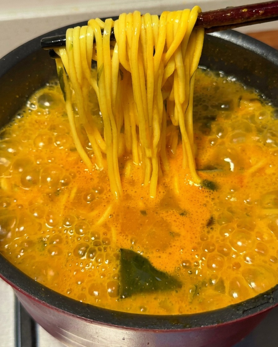 Ein Paar Essstäbchen hebt ein Bündel fertig gekochte gelbe Ramen-Nudeln aus der sprudelnden, würzigen Brühe.