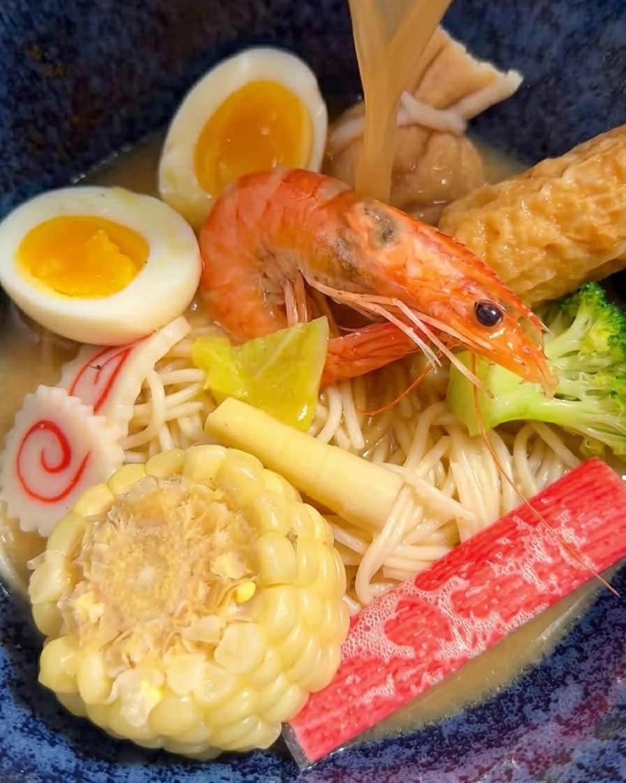 Heiße Miso-Brühe wird mit einer Schöpfkelle in eine Schüssel mit Ramen, Fleisch und Gemüse gegossen.