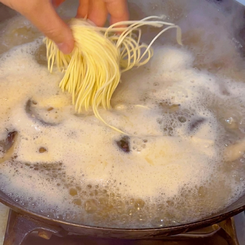 Eine Hand lässt vorsichtig ein Bündel frischer, gelber Ramen-Nudeln in die kochende Meeresfrüchtebrühe gleiten.