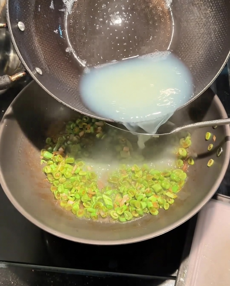 Weiße Muschelbrühe wird aus einem Topf in den Wok mit den angebratenen Bohnen gegossen.