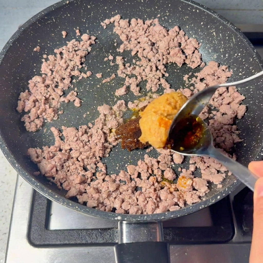 Ein großer Löffel dicke braune Paste wird in das gebratene Rinderhackfleisch eingerührt.