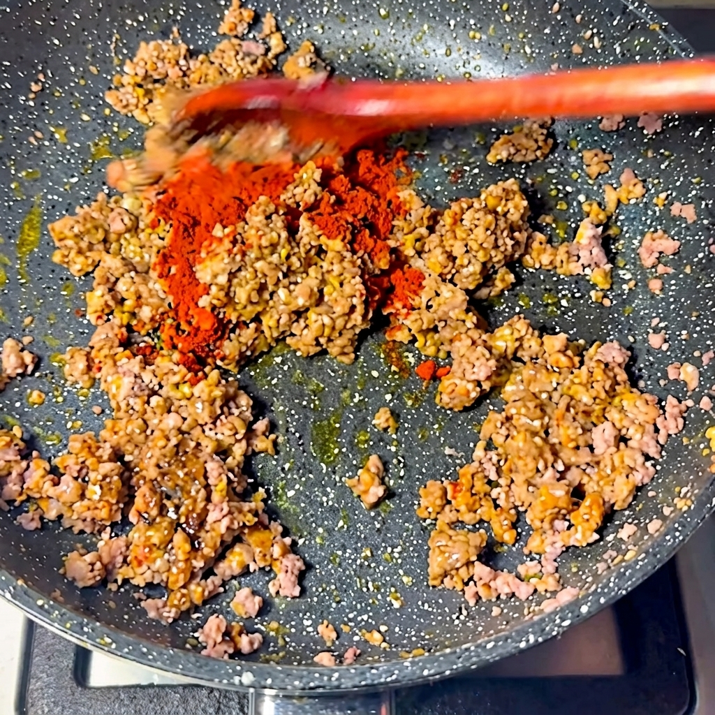 Hellrotes Chilipulver wird über die Fleischmischung in der Pfanne gestreut.