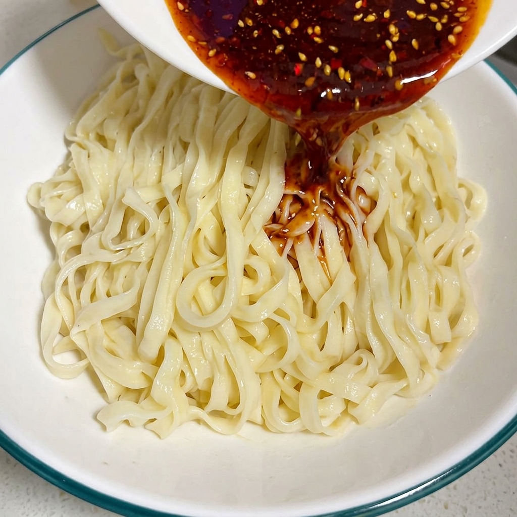 Reiche, dunkelrote Chili- und Currysauce wird aus einer kleinen Schüssel über ein Bett aus gekochten flachen Nudeln in einer größeren Schüssel gegossen.