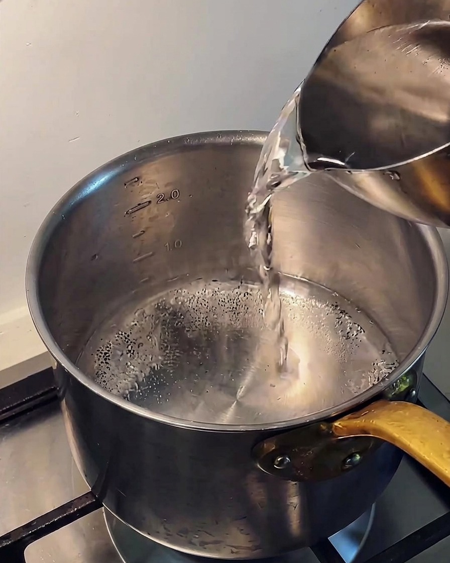 Gießen von klarem, kaltem Wasser aus einem Metallkrug in einen Edelstahltopf auf einem Herd.