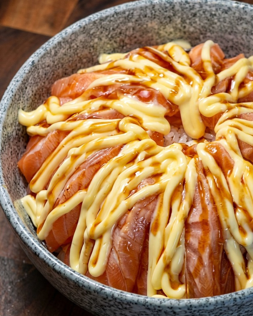 Eine Lachs-Bowl, die über der Mayonnaiseschicht mit dunkler Teriyaki-Sauce beträufelt wird.