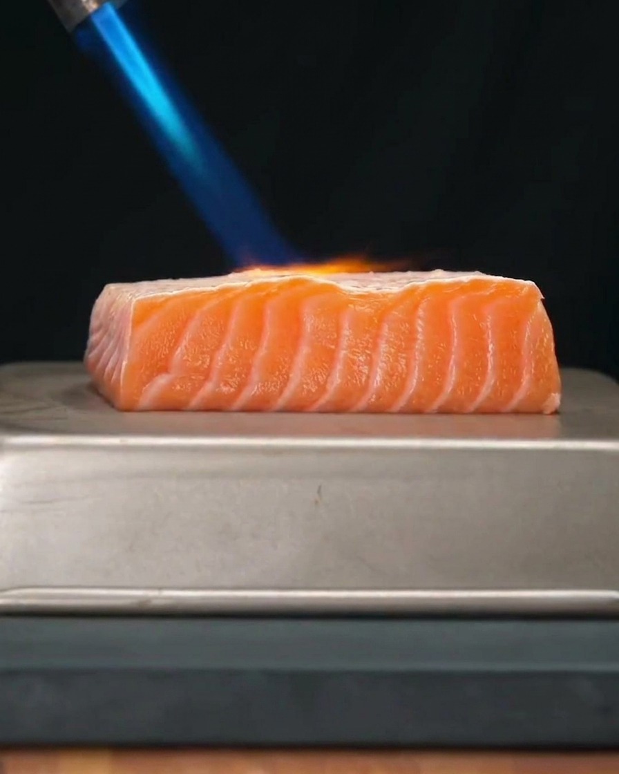Ein dickes, frisches Lachsfilet wird mit der hellblauen Flamme eines Küchenbrenners abgeflämmt.