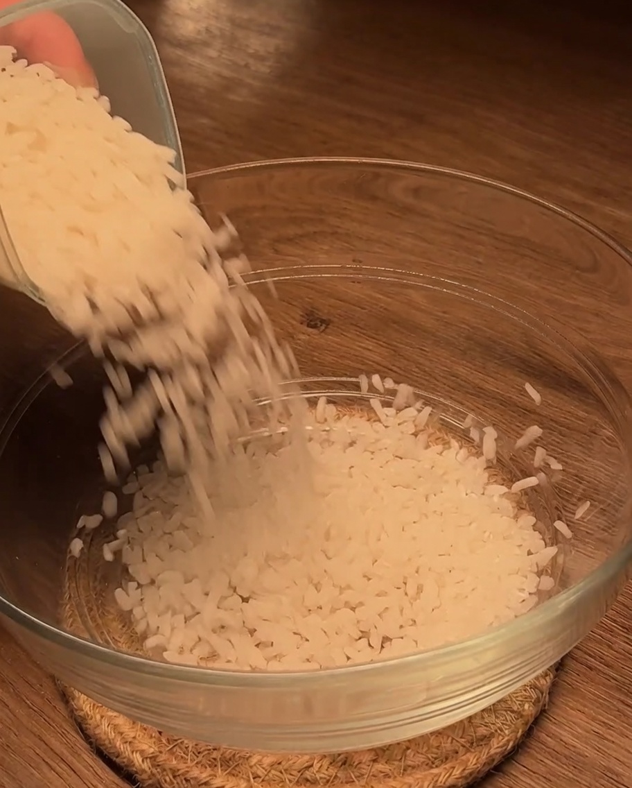 Ungekochter weißer Reis wird auf einer Holzoberfläche in eine saubere Glasschüssel gegossen.
