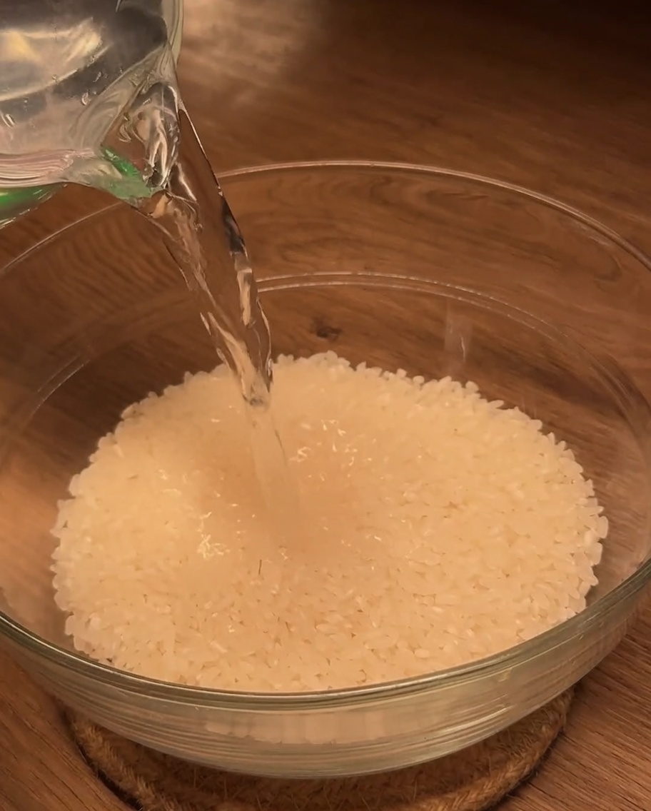 Wasser wird aus einem Messbecher in eine mit weißem Reis gefüllte Glasschüssel gegossen.