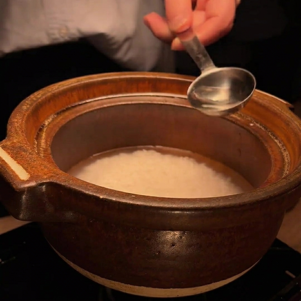 Ein Metalllöffel gießt eine kleine Menge klarer Flüssigkeit (Sake) in den Tontopf mit Reis.
