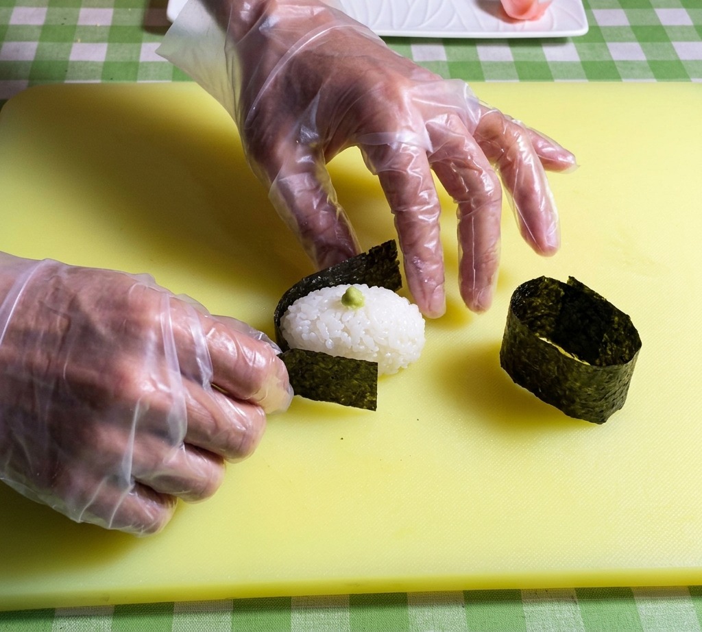 Hände platzieren einen Streifen dunkles Nori-Algenblatt um den Rand einer ovalen Sushireis-Basis.