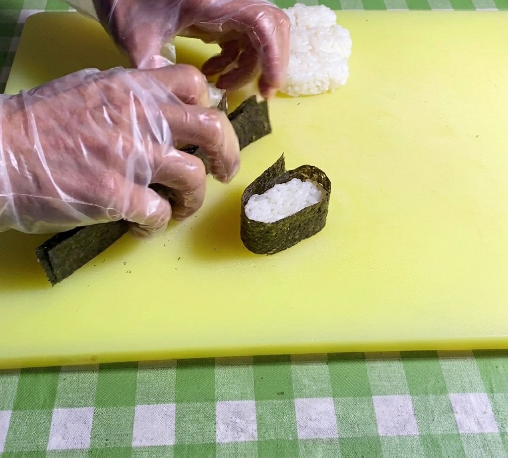 Nahaufnahme, wie Hände die Enden eines Nori-Streifens zusammendrücken, um ihn um die Reisbasis zu verschließen.