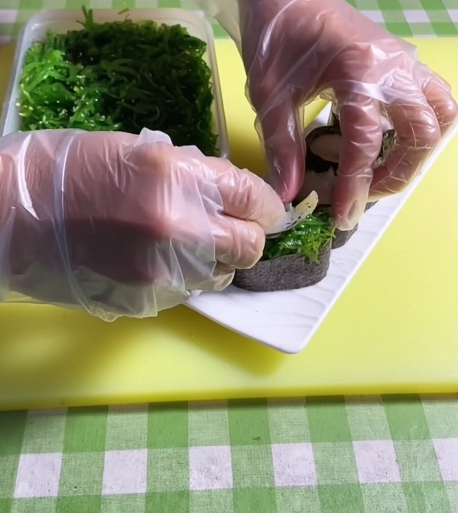 Mit einer Pinzette wird eine Portion leuchtend grüner Algensalat auf die Mitte einer Nori-umwickelten Reisbasis gesetzt.
