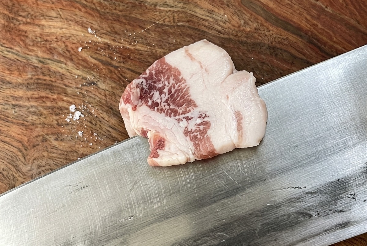Ein scharfes Messer schneidet einen rohen Schweinebauch in dünne, gleichmäßige Streifen auf einem Holzbrett.