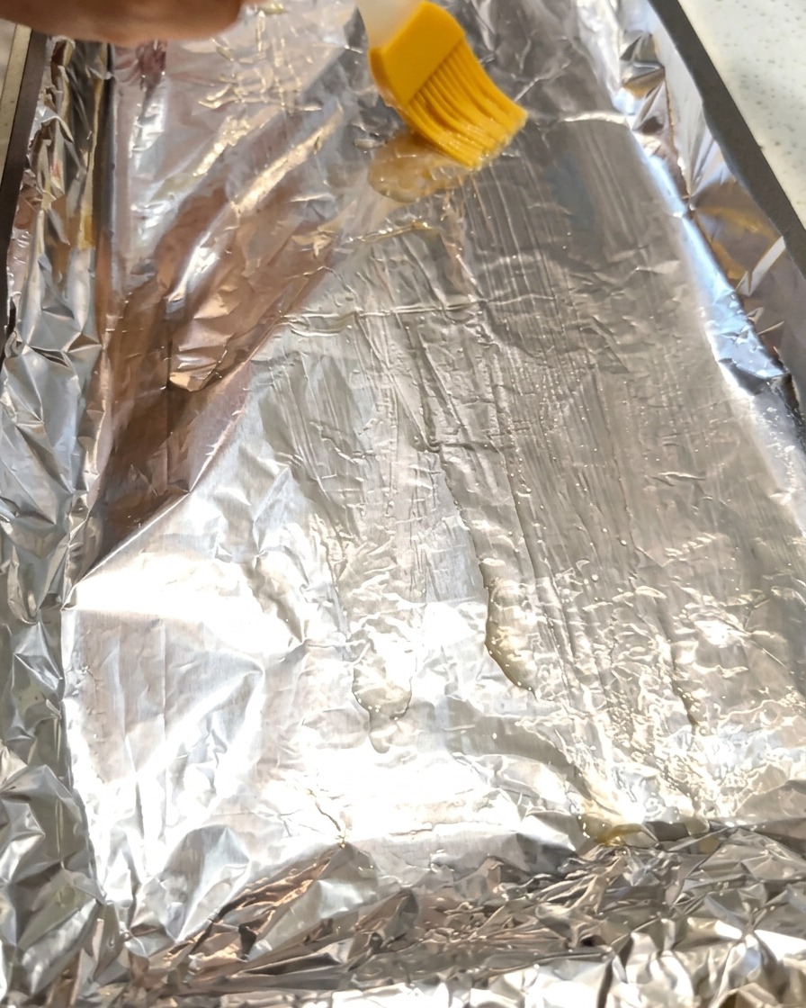 Ein Pinsel verteilt Speiseöl auf einer Alufolie auf einem Backblech.