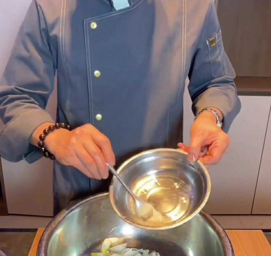 Hand gießt Natronwasser in eine Metallschüssel mit rohen Schweinebauchwürfeln, Ingwer und Frühlingszwiebelscheiben.