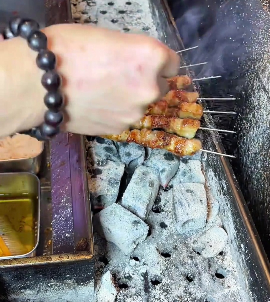 Hand streut Gewürzpulver über perfekt gegrillte, goldbraune Schweinebauchspieße auf dem Holzkohlegrill.