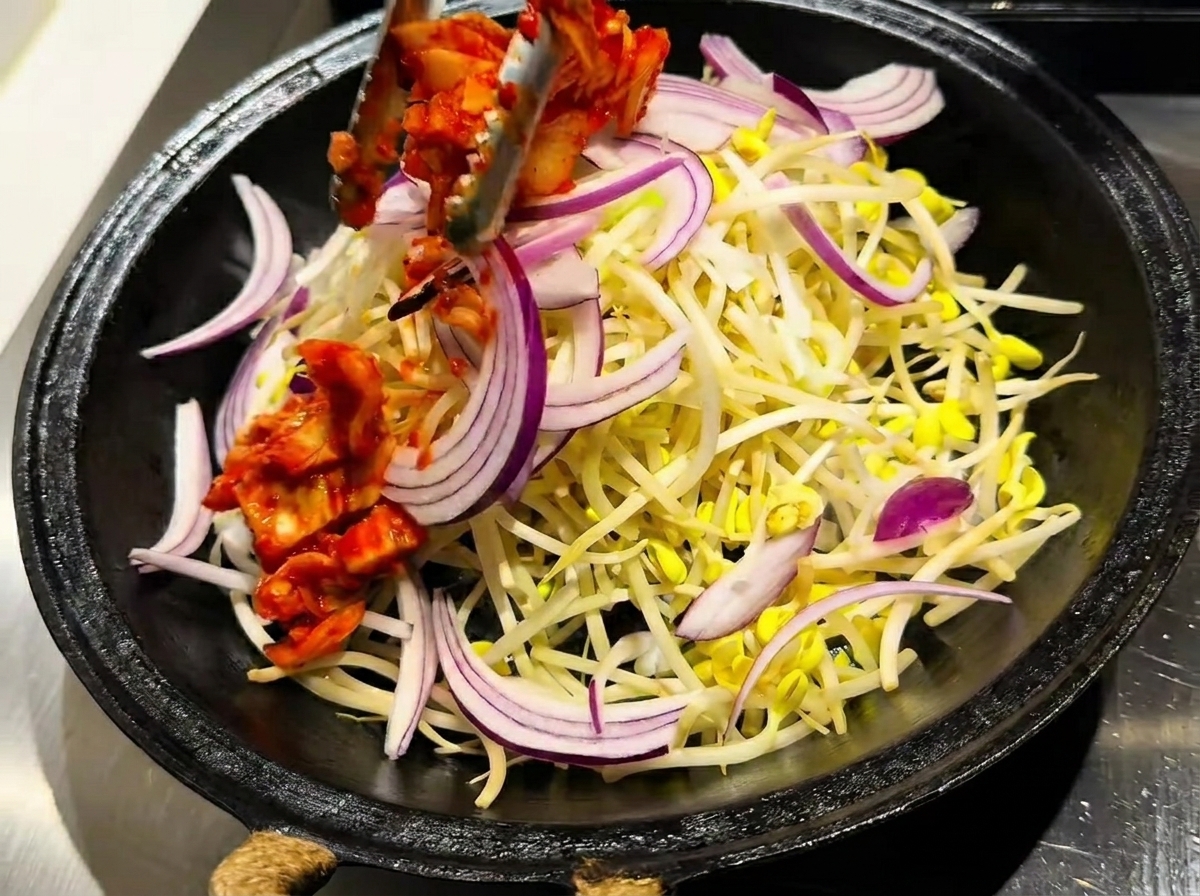 Eine Pfanne mit einer Schicht Sojasprossen, belegt mit leuchtend rotem Kimchi und rohen Zwiebelscheiben.
